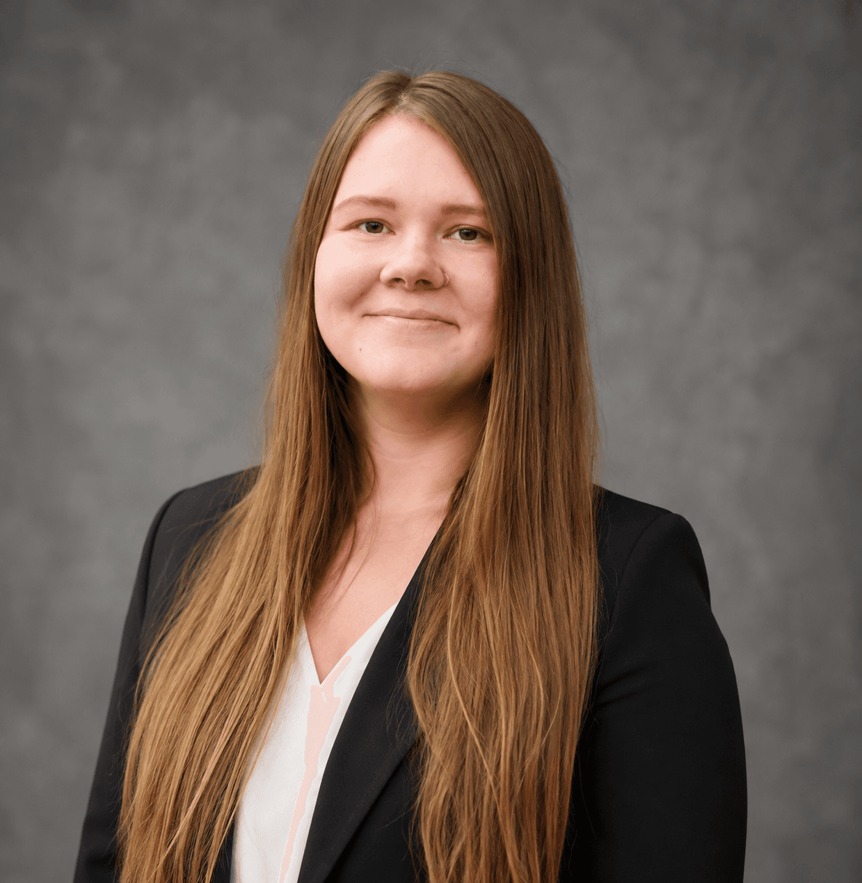 Smiling woman with long brown hair in a black blazer against a gray background.