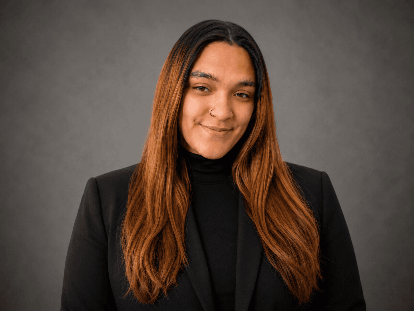 Smiling person with long auburn hair wearing a black blazer and turtleneck against grey background.