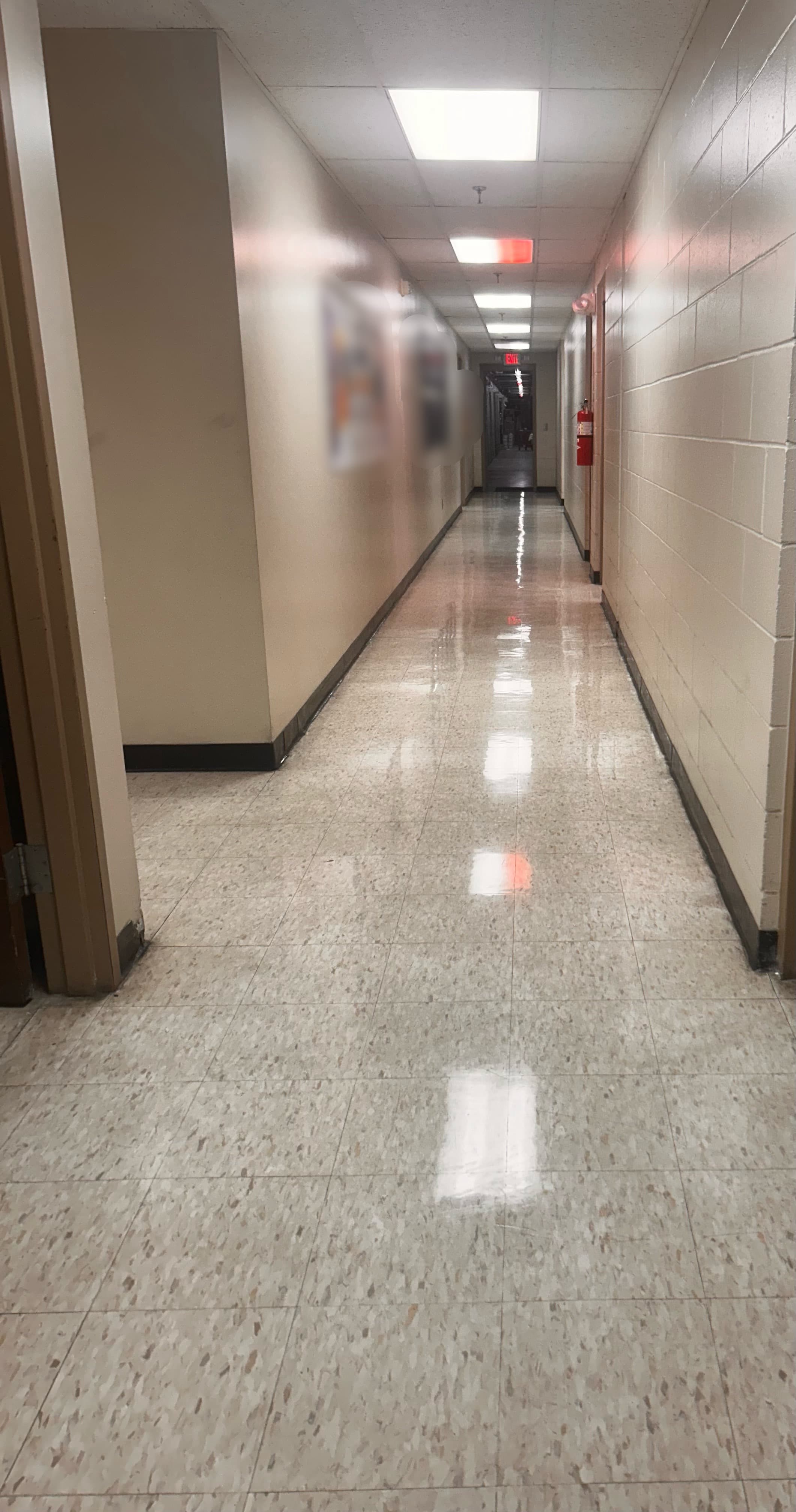 Long empty hallway with shiny tiled floors reflecting overhead lights and a red exit sign.