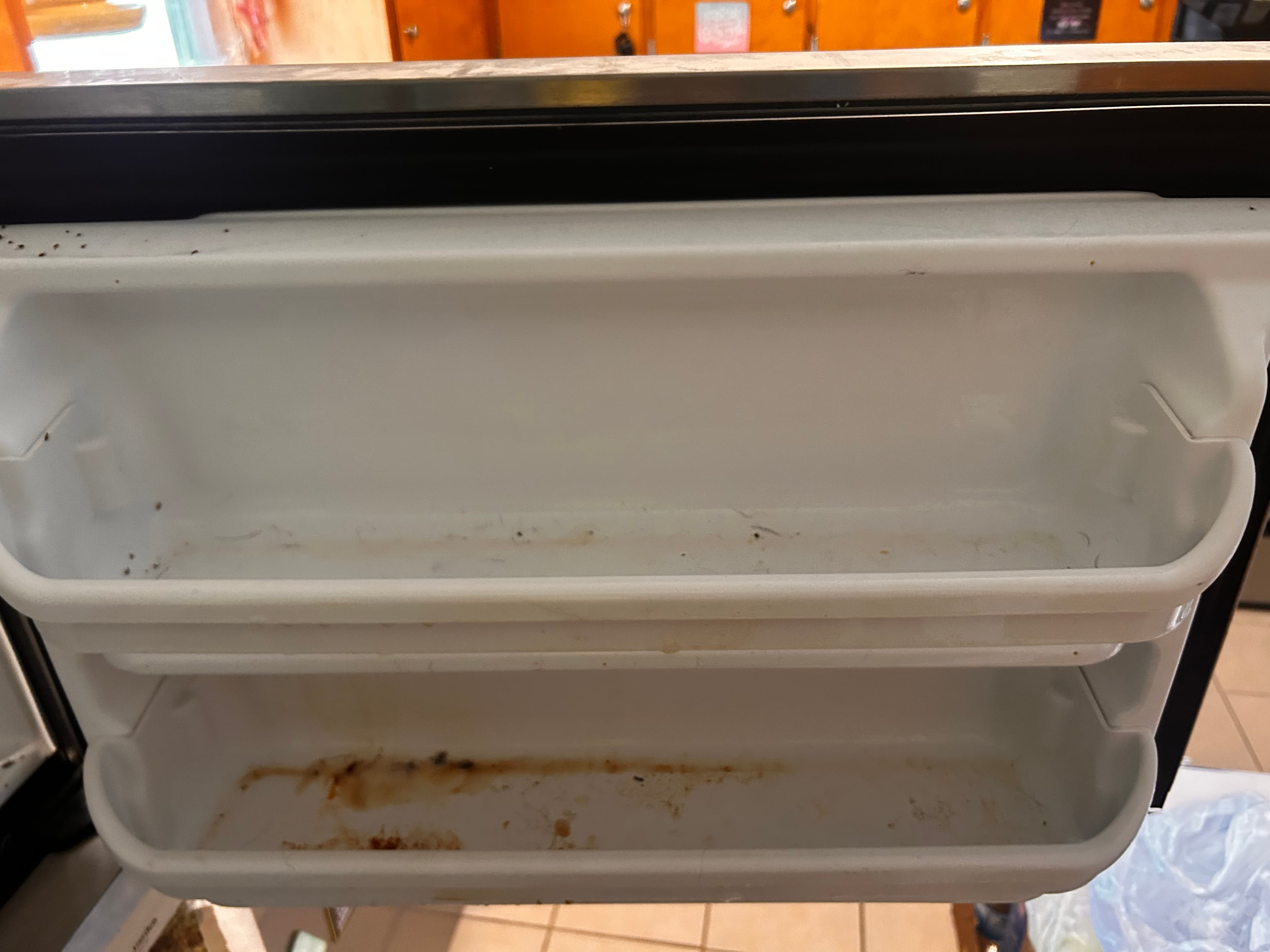 Two white plastic refrigerator door shelves covered in brown stains, crumbs, and dark debris.